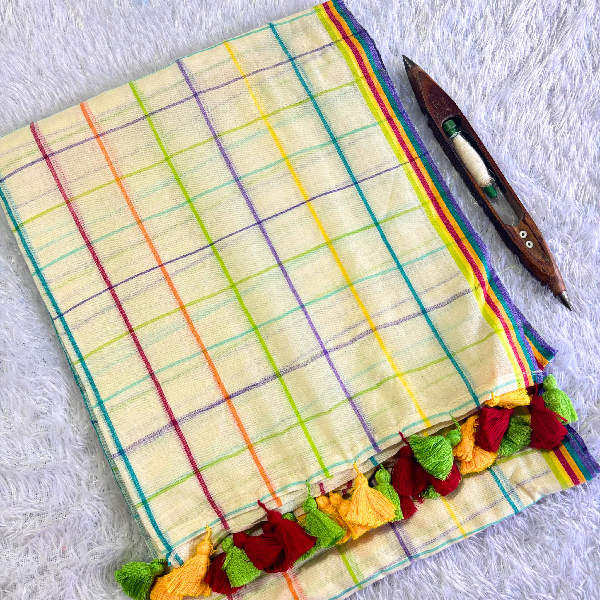 Aiswarya Lekshmi inspired cream mul cotton saree with multi-color checks and vibrant tassels next to a traditional weaving shuttle
