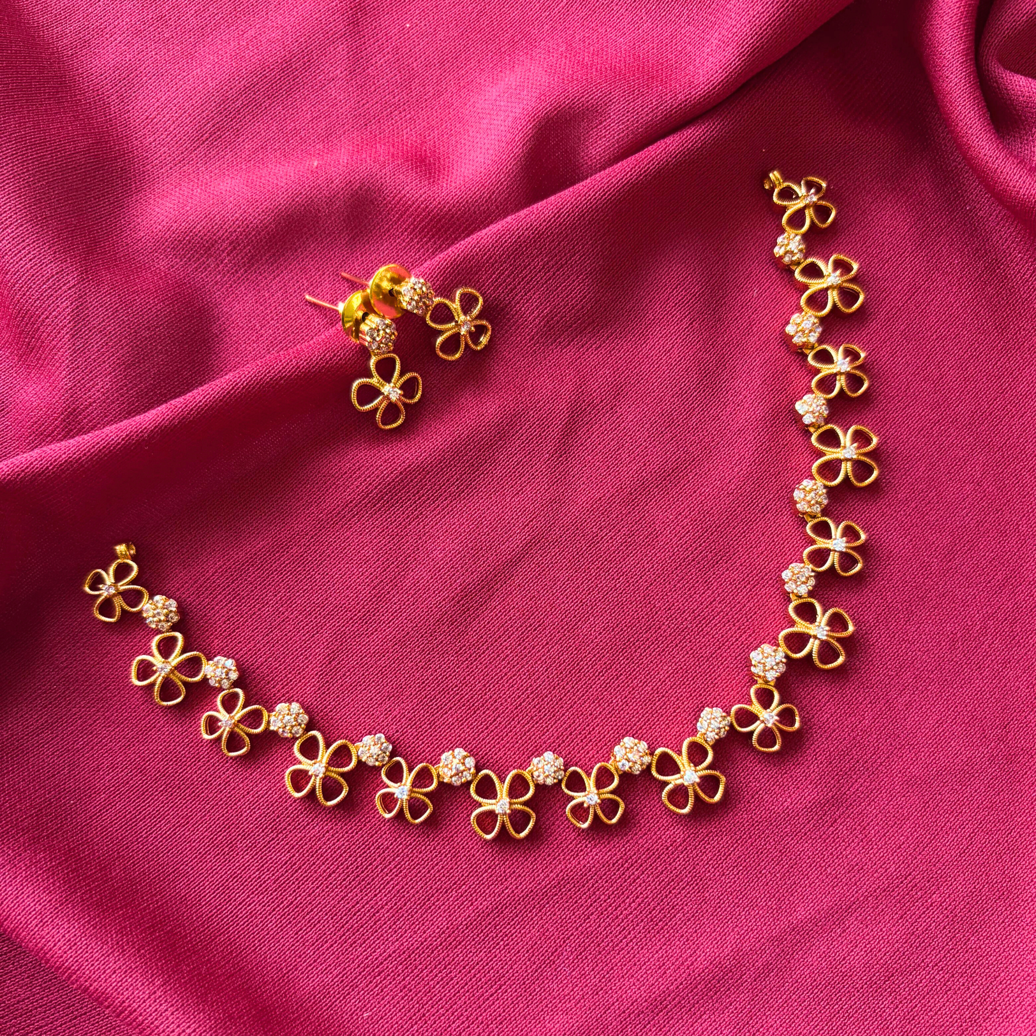 Modern gold finish necklace with four-petal floral motifs and white AD stone clusters on a dark pink fabric background